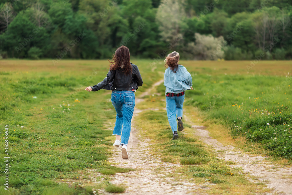 Family activity. A woman and a teen girl are joyfully running along a country road. Rear view. There is a forest in the background. The concept of freedom and happiness Family activity. A woman and a teen girl are joyfully running along a country road. Rear view. There is a forest in the background. The concept of freedom and happiness