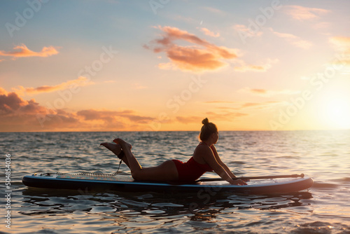 Silhouette of plus sized woman in bikini posing lying on a sup board. Copy space. Sunset sky on the background. The concept of water sports and summer vacation © _KUBE_