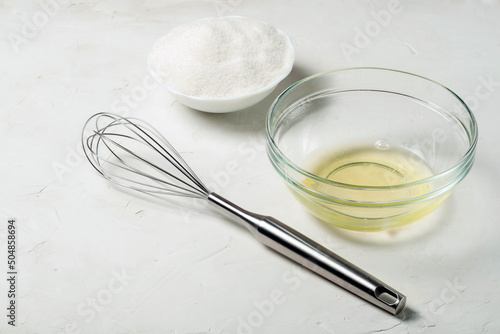 ingredients for the French meringue dessert are egg white, sugar and a whisk on a gray surface.