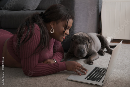 African young woman in sportswear laying on the floor using a laptop in the living room with her sharpei dog at home. photo