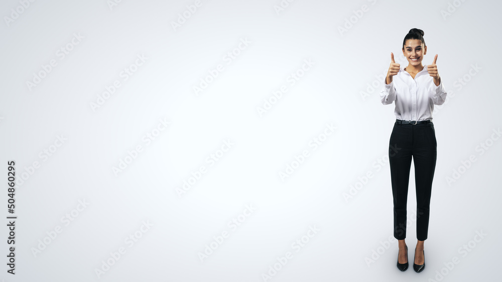 Like concept with happy young businesswoman in white shirt and black trousers holding thumbs up and standing full length on light grey background with blank space for you logo, mock up