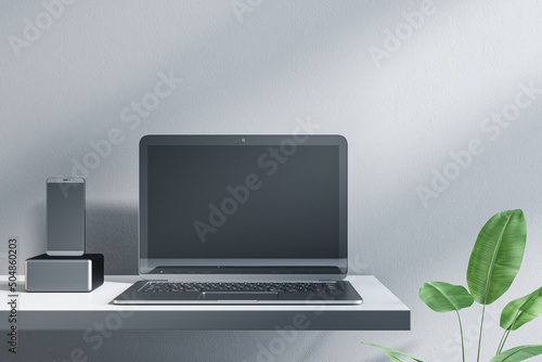 Close up of empty laptop and smartphone on designer office desktop. Decorative plant and concrete wall background. Mock up, advertisement, technology, product review and webinar concept. 3D Rendering.