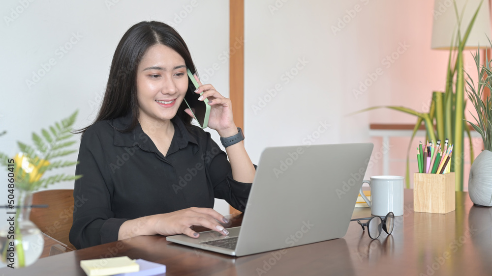 Smiling female entrepreneur working on laptop computer and having conversation on mobile phone Smiling female entrepreneur working on laptop computer and having conversation on mobile phone