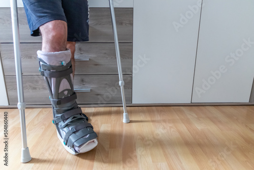 European man after Achilles tendon rupture operation is back home with moon boot special physiotherapy shoe and crutches for recovery at home against the hurting leg learning to walk first steps PWB