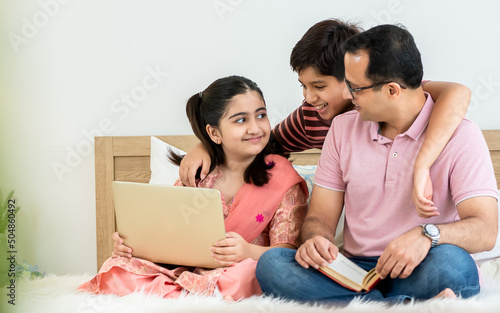 Indian single father, teenage son, daughter using laptop, sitting on bed at home in holiday, watching movie, online learning, doing homework together, happily smiling. Education, Family Concept.