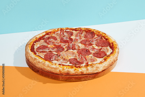 talian pizza with salami, ham, bacon, pepperoni on coloured background. Meat pizza with sausage and bacon in minimal style on blue, orange color. American pizza delivery concept with color backdrop. photo