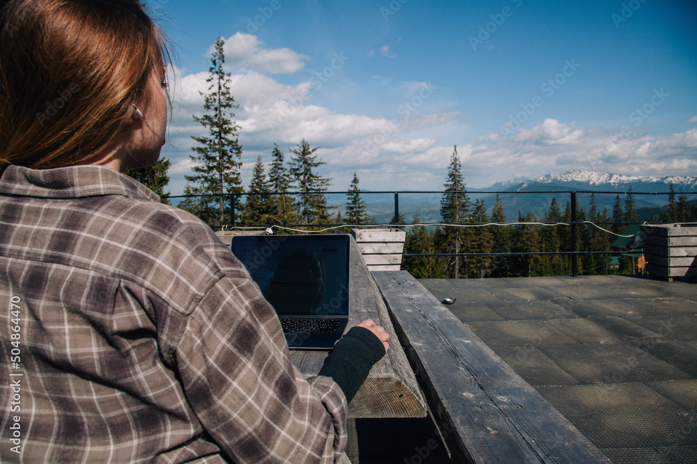 A young, red-haired girl in a plaid shirt sits at an old wooden table in nature against the backdrop of mountains and works on a laptop. typing. view from the back.