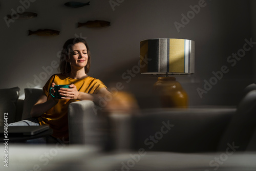 Smiling woman with eyes closed holding coffee mug sitting in living room photo