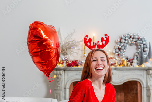 Happy woman wearing Christmas headdress by red heart shape balloon at home