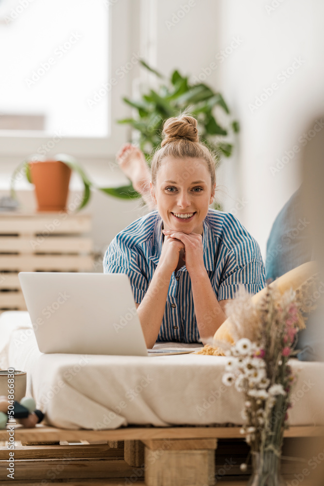 Smiling businesswoman with hand on chin lying by laptop on bed at home Smiling businesswoman with hand on chin lying by laptop on bed at home