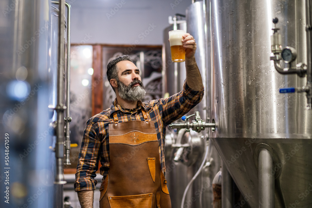 Bearded brewery master holding glass of beer and evaluating its visual characteristics. Small family business, production of craft beer. - obrazy, fototapety, plakaty 