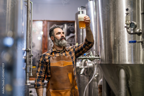Bearded brewery master holding glass of beer and evaluating its visual characteristics. Small family business, production of craft beer. photo