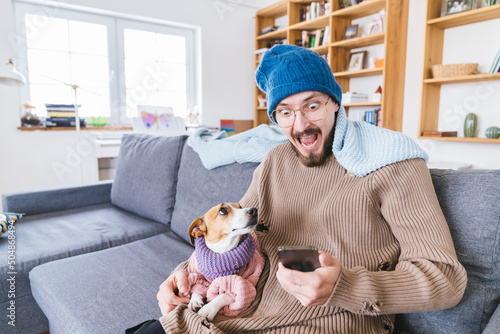 Shocked man with wooly hat sitting on couch holding dog checking smartphone photo