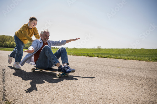 Playful granddaughter pushing happy grandfather sitting on skateboard