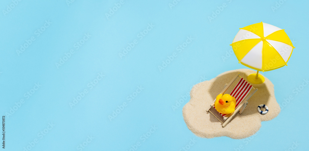 Fototapeta premium Summer concept. Sun umbrella and duck in sun lounger on sea sand on blue background. Sun protection concept in summer.