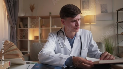 Junior doctor. Positive man. Online consultation. Smiling guy in medical clothes talking on camera with x-ray photo of lungs making reference to science book sitting desk light room interior. photo