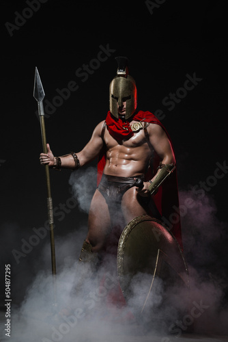 A young athletic man dressed as a Roman soldier in a red cloak stands with a spear in hand and a full-length shield on a black background, shrouded in mysterious smoke. photo