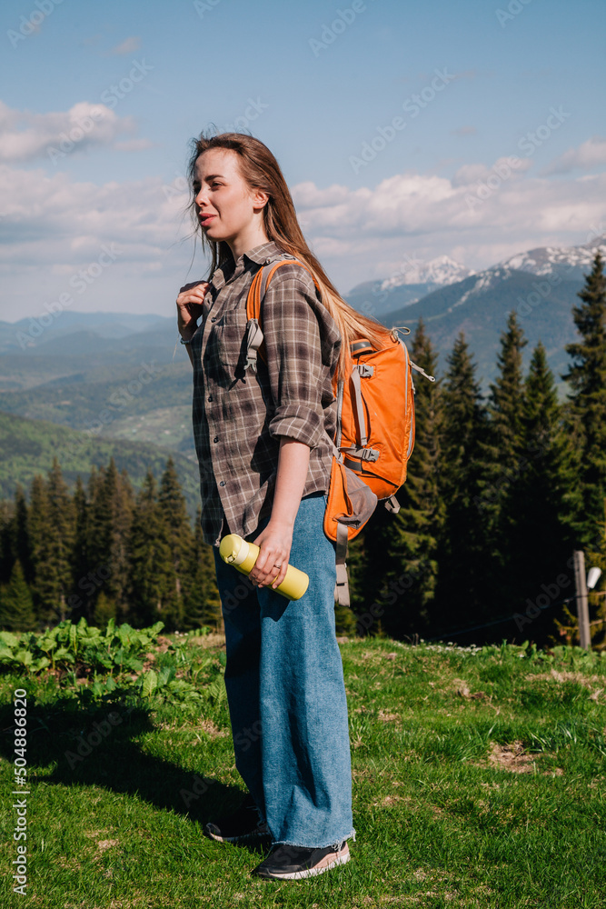 A young, girl in a plaid shirt and jeans with loose hair with an orange backpack drinks tea from a bright thermal cup against the backdrop of the mountains in the Carpathians. view from the back.