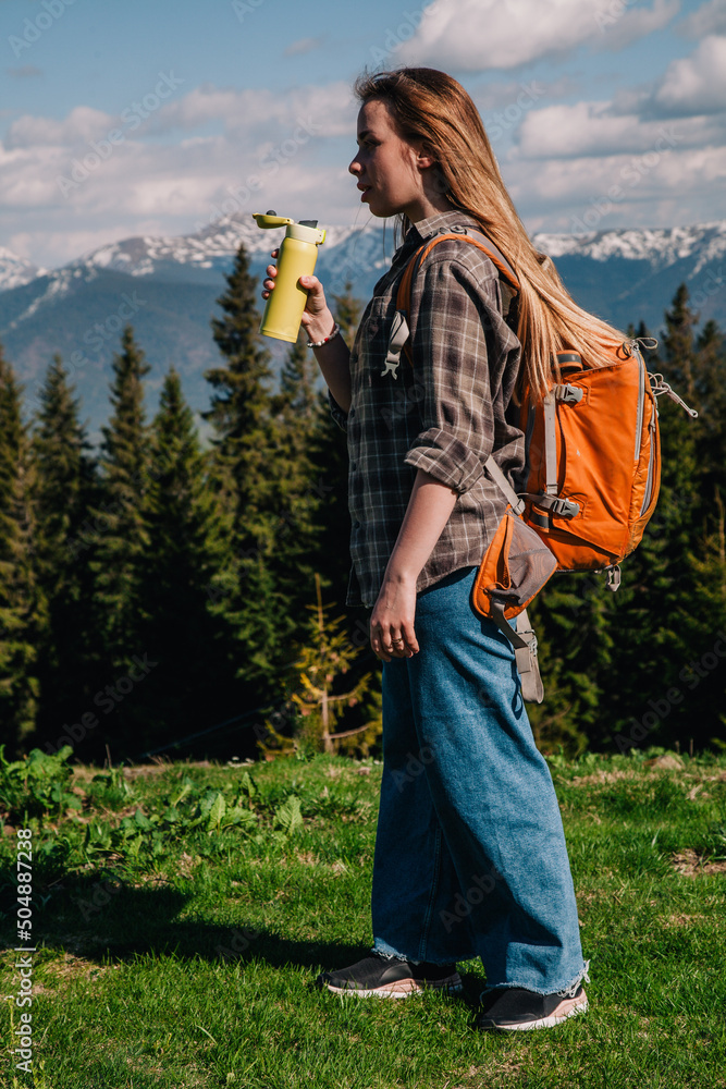 Naklejka premium A young, girl in a plaid shirt and jeans with loose hair with an orange backpack drinks tea from a bright thermal cup against the backdrop of the mountains in the Carpathians. view from the back.