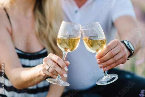 Man and woman hold glasses with white wine on background of a lavender field. Young couple drink rose wine in sunset lavender field. Romantic evening in sunset. Summer in Provence, France. Closeup.