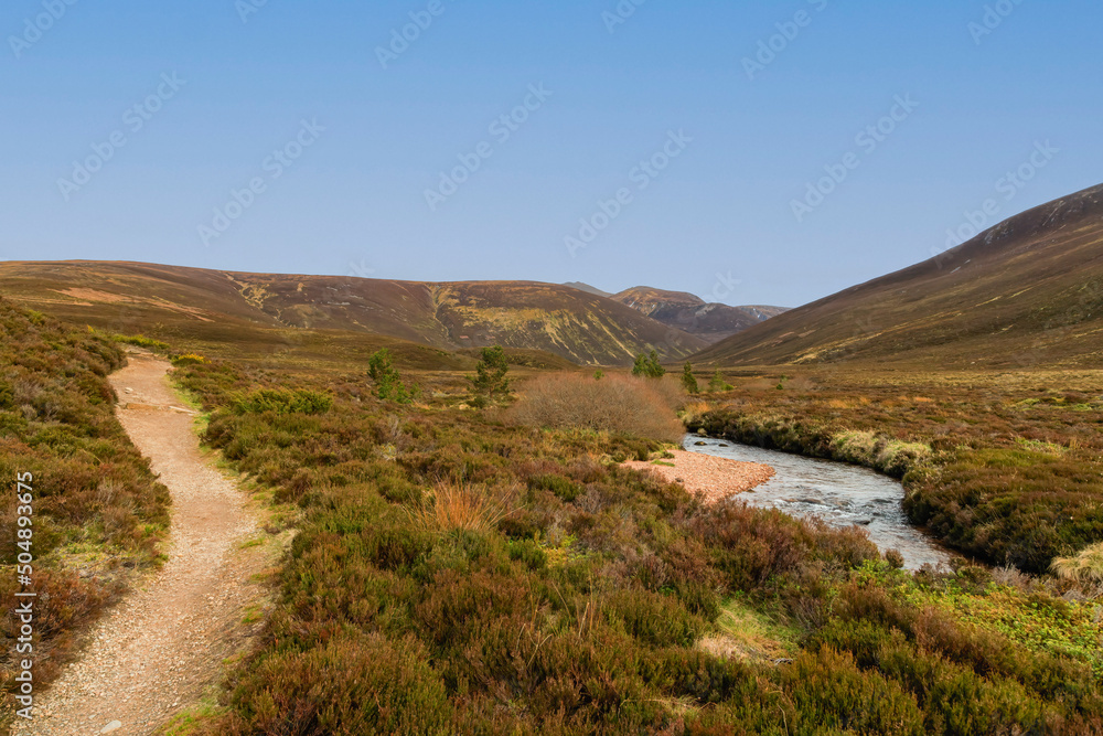 A scenic view of a Scottish moutain river (Nethy) with moorland and path in the foreground and mountain valley and summit in the background under a beautiful blue sky