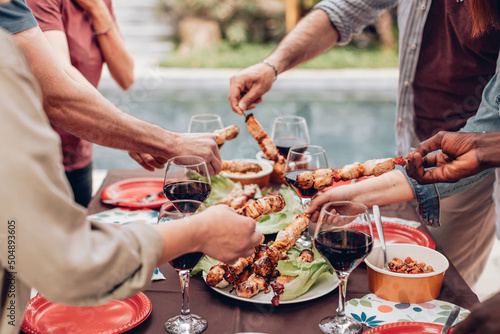 hands grabbing skewers from the table with wine glasses at the poolside summer barbecue party