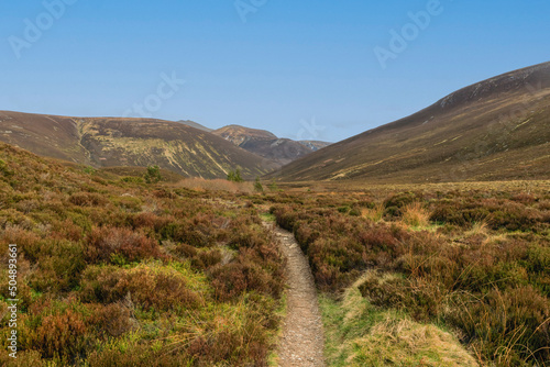 A scenic view of a Scottish moutain path with moorland in the foreground and mountain valley and summit in the background under a beautiful blue sky