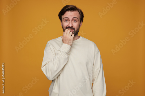 indoor portrait of man wears white sweatshirt posing over yellow background biting his nails and feels guilty