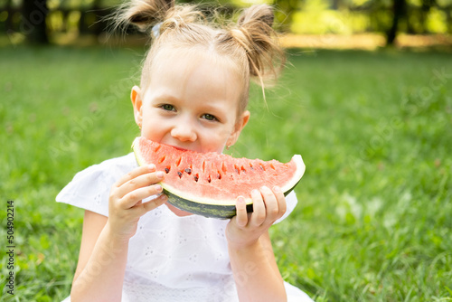 funny smiling kid girl in white dress eating watermelon at green lawn