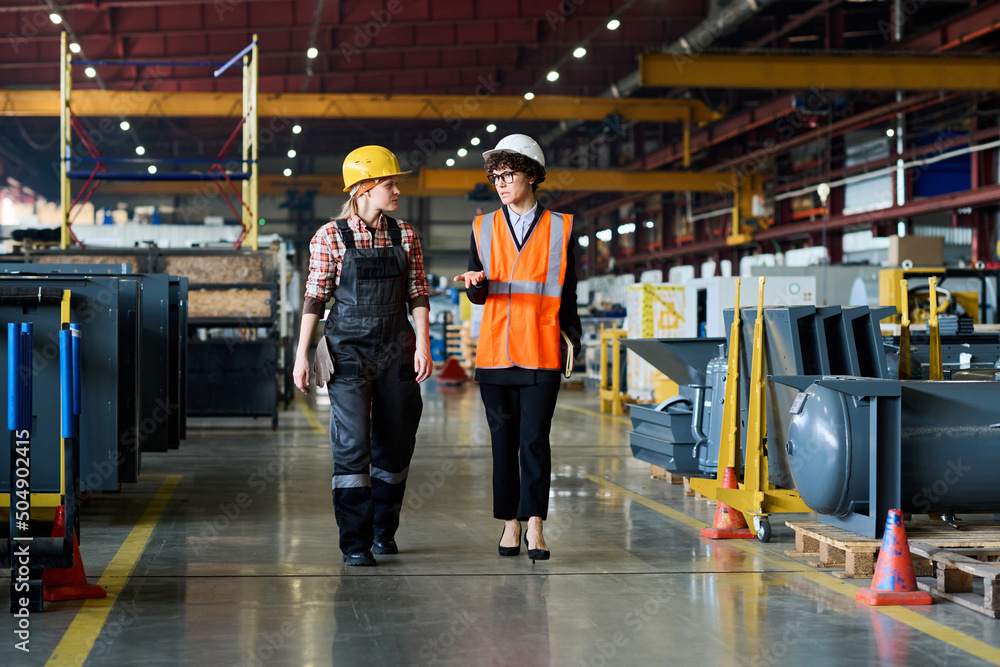 Fototapeta premium Two young workers of modern industrial factory having discussion of work while moving along aisle between new equipment
