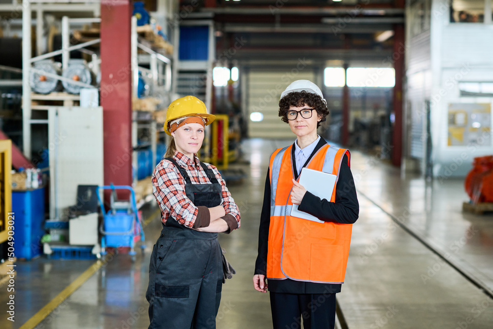 Two young confident female colleagues in workwear looking at camera while standing in workshop against new industrial equipment