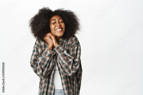 Image of lovely and cute african woman dreaming of something silly, smiling with eyes closed, daydreaming, standing happy against white background © Cookie Studio