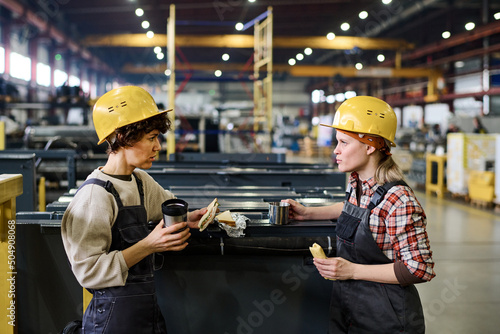 Two young engineers or workers of warehouse having sandwiches and hot tea at lunch break and discussing latest news or working points