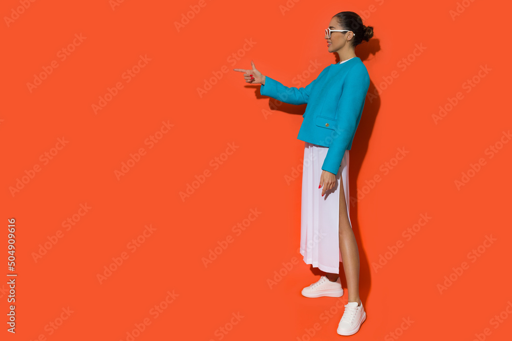 Confident young woman in blue jacket, white dress with slit and sneakers is pointing to the side. Confident young woman in blue jacket, white dress with slit and sneakers is pointing to the side.