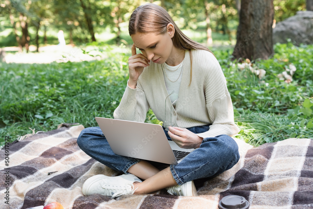 freelancer sitting on blanket with crossed legs and using laptop while holding glasses. freelancer sitting on blanket with crossed legs and using laptop while holding glasses.