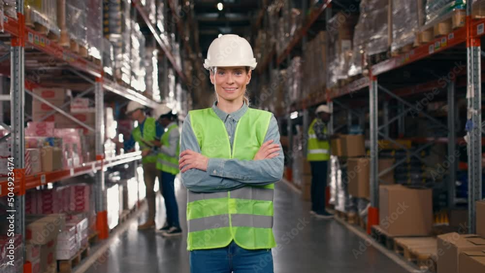 Portrait of cheerful Caucasian young woman employee in helmet standing in big warehouse looking at camera and smiling in good mood. Storage, cardboard boxes with goods, storehouse, work concept Portrait of cheerful Caucasian young woman employee in helmet standing in big warehouse looking at camera and smiling in good mood. Storage, cardboard boxes with goods, storehouse, work concept
