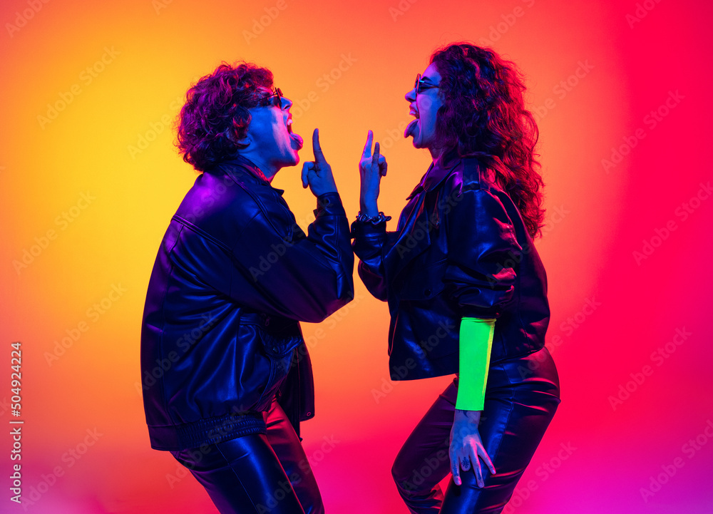 Fototapeta premium Excited young man and girl in black leather outfits having fun, gesturing, shouting and shaking head over yellow-red background in neon light