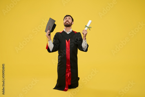 Frint view of brunette student wearing graduate gown standing on knees. Young male holding diploma and mortarboard, looking up, happy, glad, grateful. Concept of education. photo