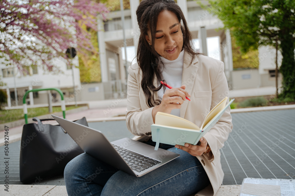 Obraz premium Smiling university student studying taking notes using laptop computer. Young freelancer working outdoors sitting at workplace 