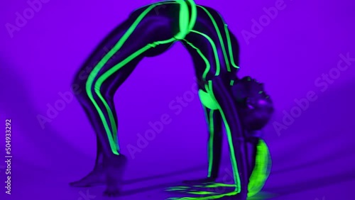 Futuristic flexible woman in neon costume doing forward somersault bending looking at camera in ultraviolet light. Wide shot slim gymnast performing modern dance posing photo