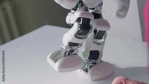 A white robot dancer with luminous blue eyes calmly raises its leg and makes movements with its arms and stands stably on one leg, keeping its balance. Closeup photo