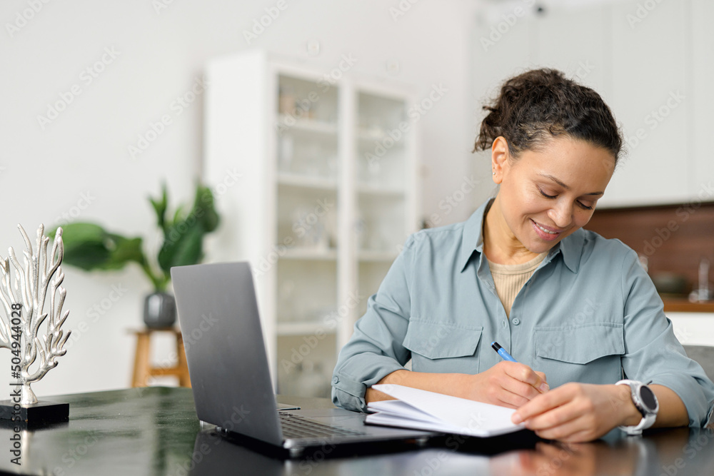 Woman is using laptop computer for remote work or studying online, she takes notes watching webinars or classes at kitchen table at home Woman is using laptop computer for remote work or studying online, she takes notes watching webinars or classes at kitchen table at home