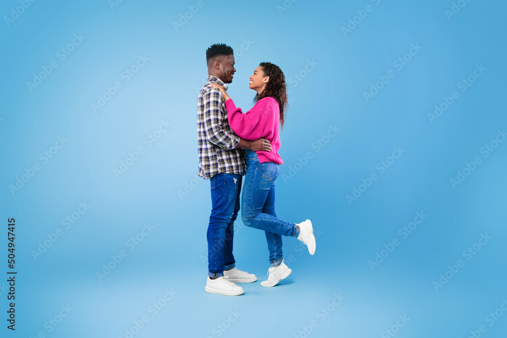 Side view of happy young African American couple in casual wear hugging and looking at each other on blue background Side view of happy young African American couple in casual wear hugging and looking at each other on blue background