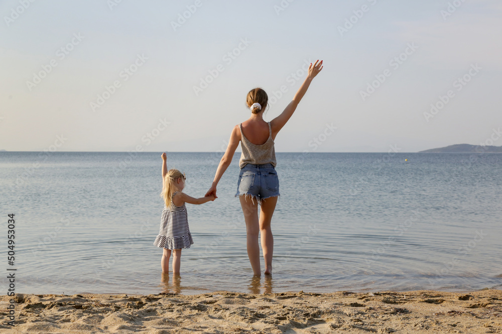 Mother and daughter on the beach looking to horizon over the sea with hands raised up. Family summer vacation concept.	