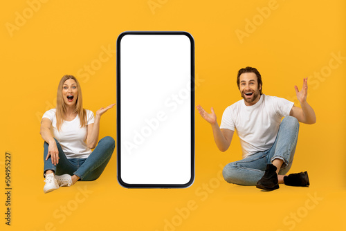 Cheerful surprised young european man and lady with open mouths raise hands, sit on floor with huge phone