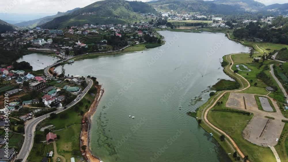 Several boats and watercraft navigate in the blue reservoir Gregory Lake while several vehicles drive left-hand on the winding provincial road between the mountains in Sri Lanka. Drone panning shot Several boats and watercraft navigate in the blue reservoir Gregory Lake while several vehicles drive left-hand on the winding provincial road between the mountains in Sri Lanka. Drone panning shot