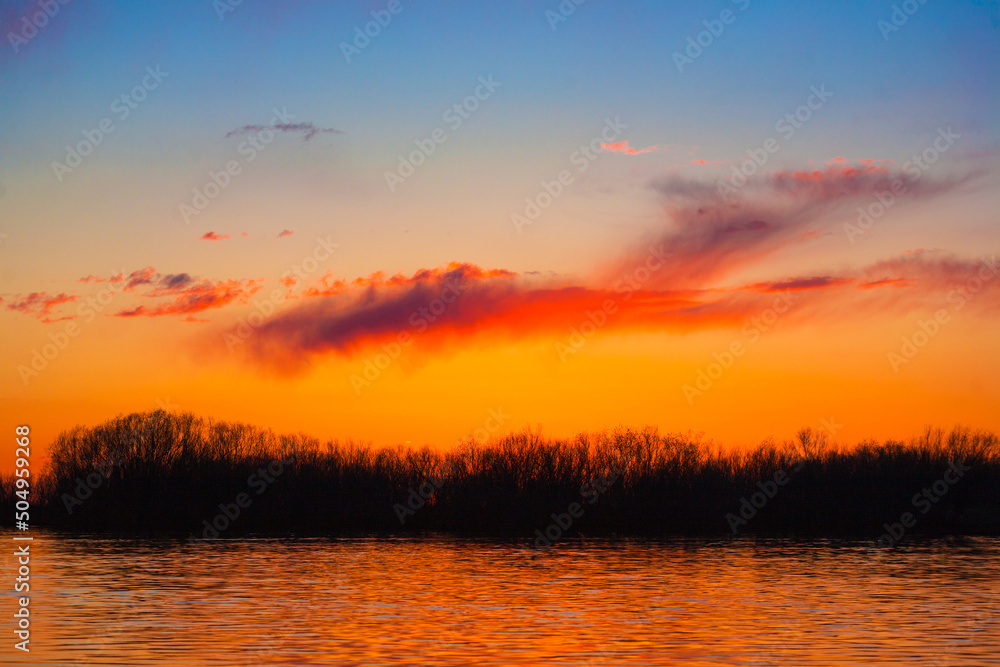 Orange, yellow and purple sunset on river with dark colorful clouds in sky with trees reflection in water