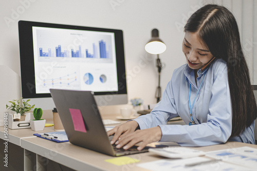 Asian female employee talking on the phone and working in a laptop with a smiling face, Office staff work and telephone conversations.