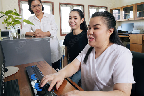 Wide angle view of happy Asian women co-workers in office workplace including person with blindness disability using computer with refreshable braille display assistive device. Disability inclusion.
