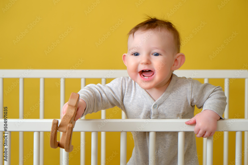 Happy toddler baby boy in a crib with a wooden toy in his hand, yellow studio background. A smiling child in pajamas Happy toddler baby boy in a crib with a wooden toy in his hand, yellow studio background. A smiling child in pajamas